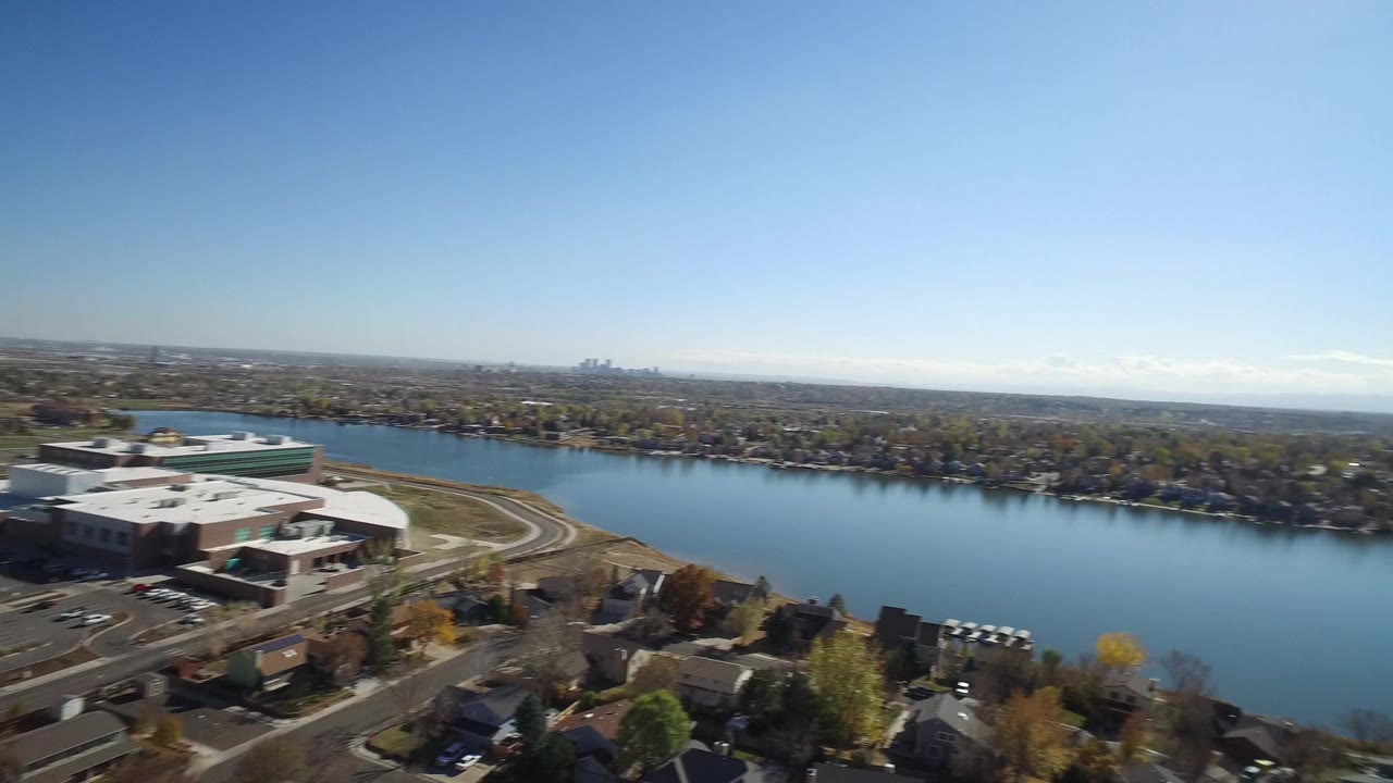 Early aerial frame in Hidden Lake, Colorado with the lake across the center, a large building complex at left, homes along the shore, distant skyline, and no visible people.