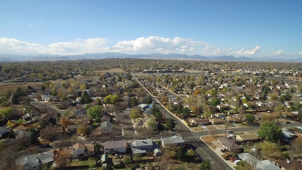 Aerial view over residential streets, houses, trees, blue sky, and distant mountains.