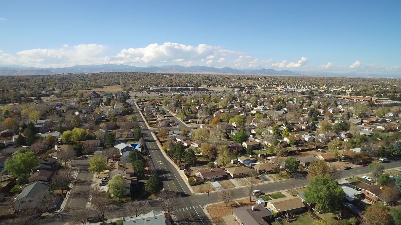 Opening aerial frame over Hidden Lake Colorado homes and tree-lined streets, with a long road at left, distant mountains, scattered clouds, and no visible people.
