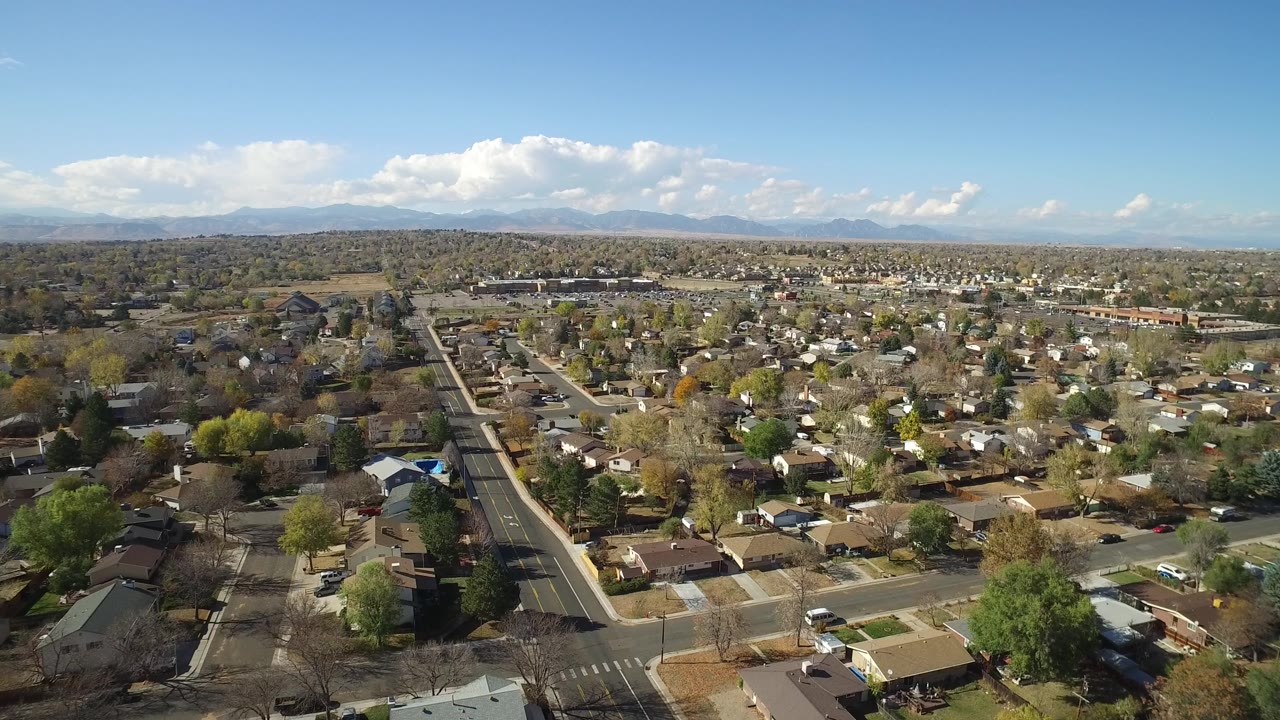 Aerial frame over Hidden Lake Colorado with rooftops spreading across the center, a straight street running toward the horizon, pale mountains, cloud bands, and no visible people.