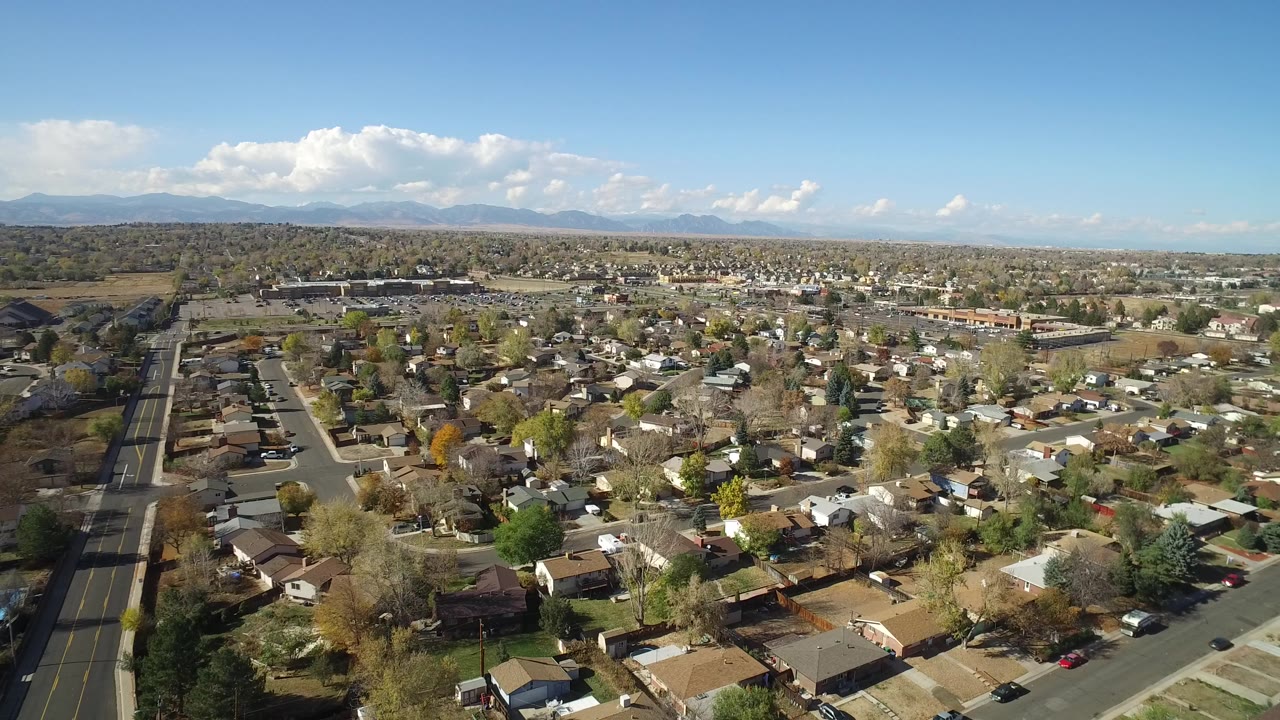 Aerial view over residential streets, homes, trees, distant hills, and blue sky.