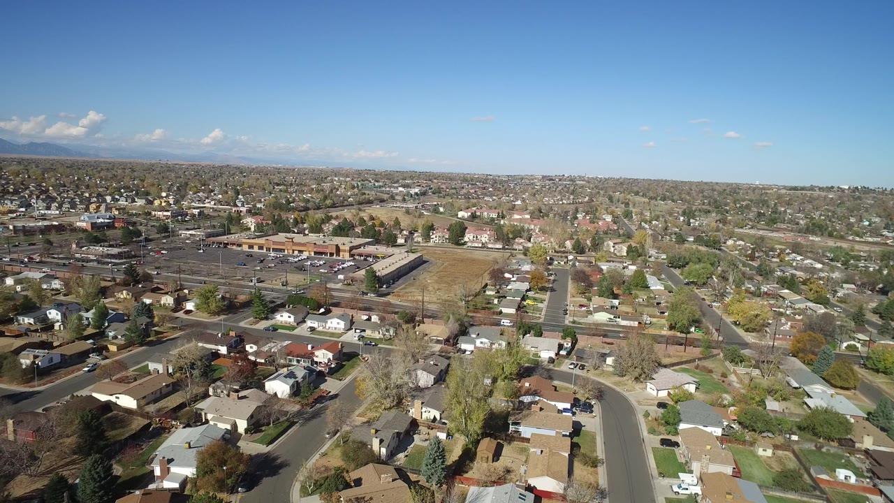 Aerial view over homes, streets, parking lots, and distant hills under blue sky.