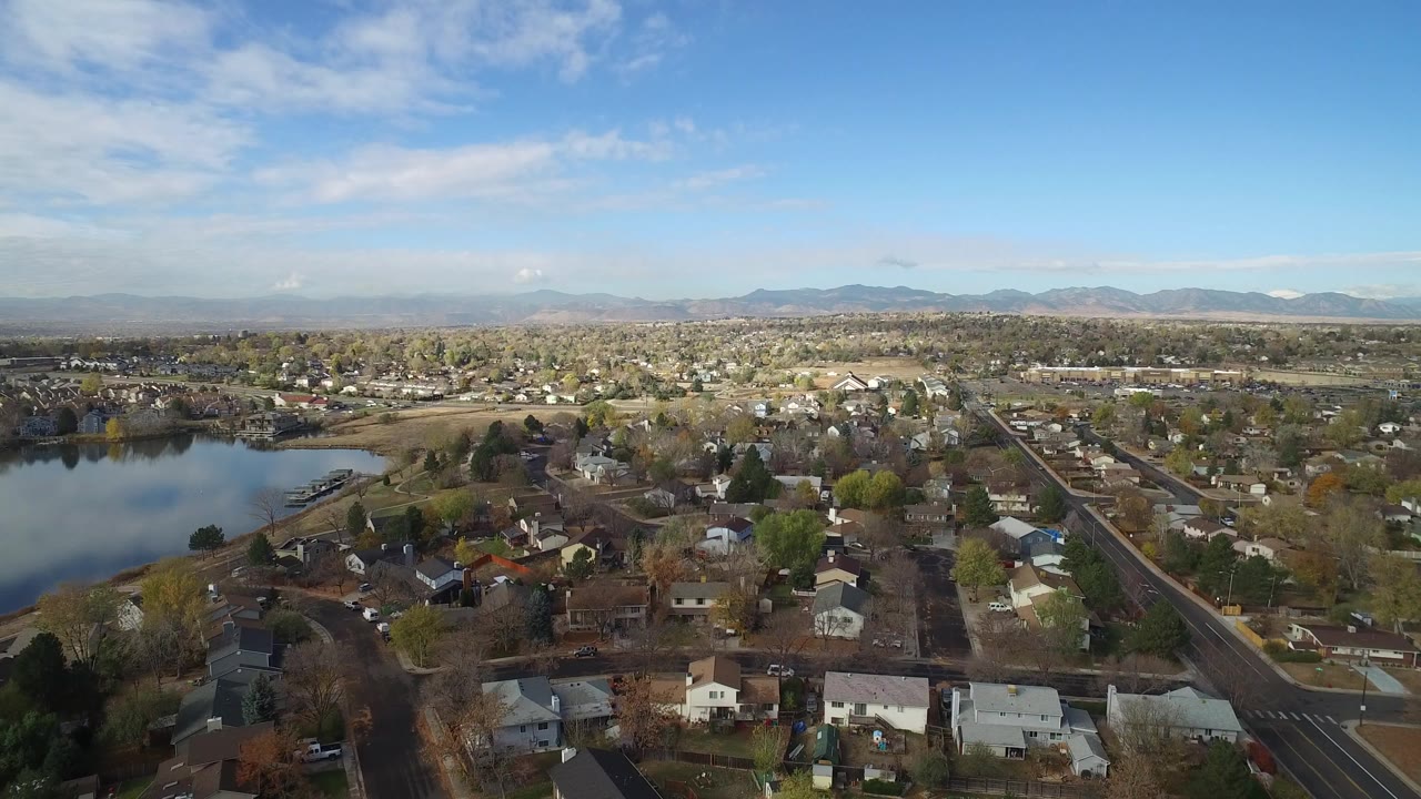 Opening aerial frame over Hidden Lake, Colorado, with a blue lake on the left, residential houses below, a road to the right, distant mountains on the horizon, and no visible people.