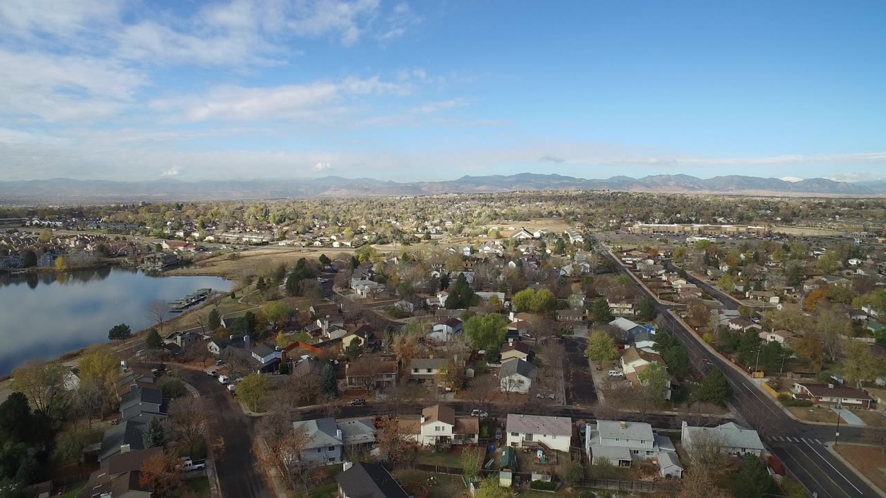 Wide aerial over residential blocks beside a small lake under blue sky.