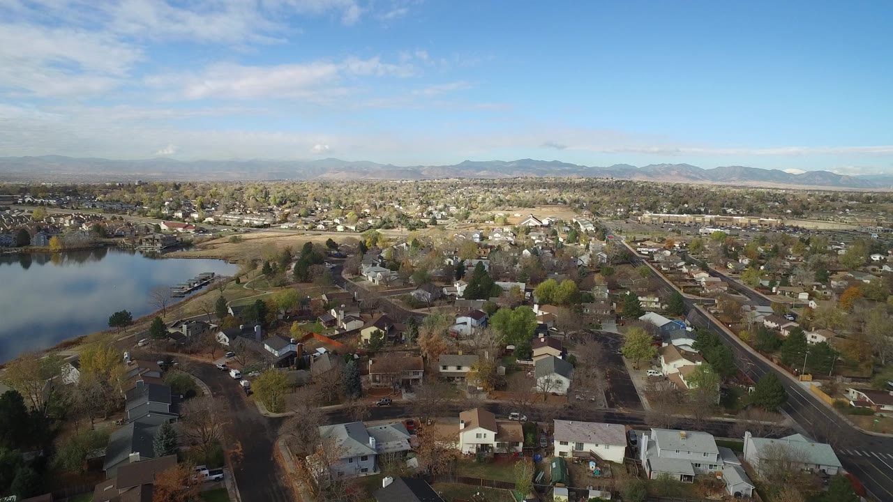 Wide aerial view over Hidden Lake, Colorado homes with the lake at left, rooftops in the foreground, distant mountains, blue sky, and no visible people.