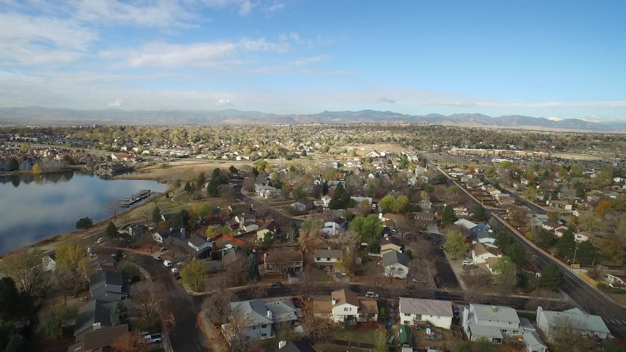 Residential aerial with rooftops, streets, lake, and mountain backdrop.