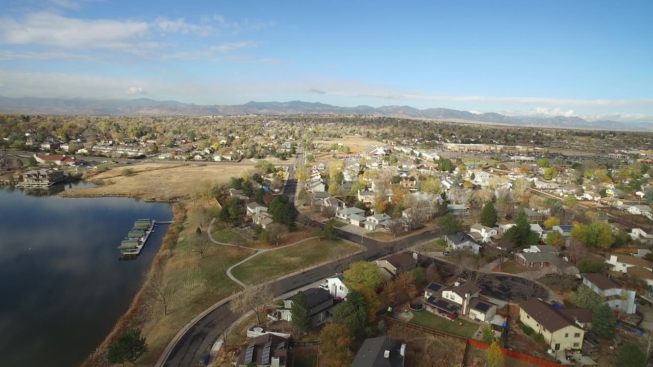 Opening aerial frame over Hidden Lake, Colorado with dark water at left, docks near the shore, residential streets and houses to the right, distant mountains, and no visible people.