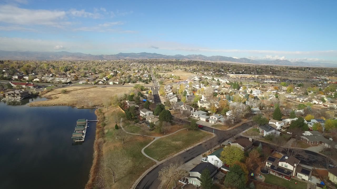 Opening aerial frame over Hidden Lake, Colorado, with dark lake water and docks at left, homes and curving streets in the foreground, a broad residential area beyond, distant mountains, and no visible people.