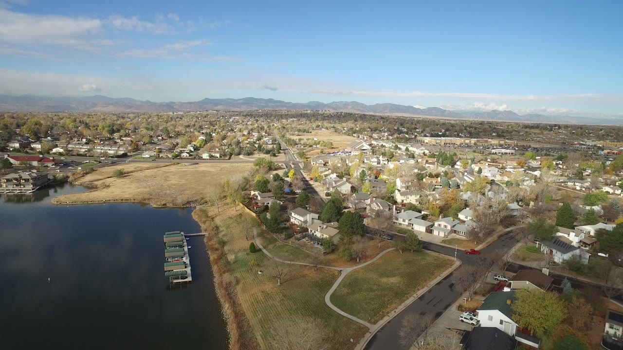 Opening aerial frame over Hidden Lake, Colorado, with dark water and small docks at left, curved paths and homes at right, distant mountains on the horizon, and no visible people.