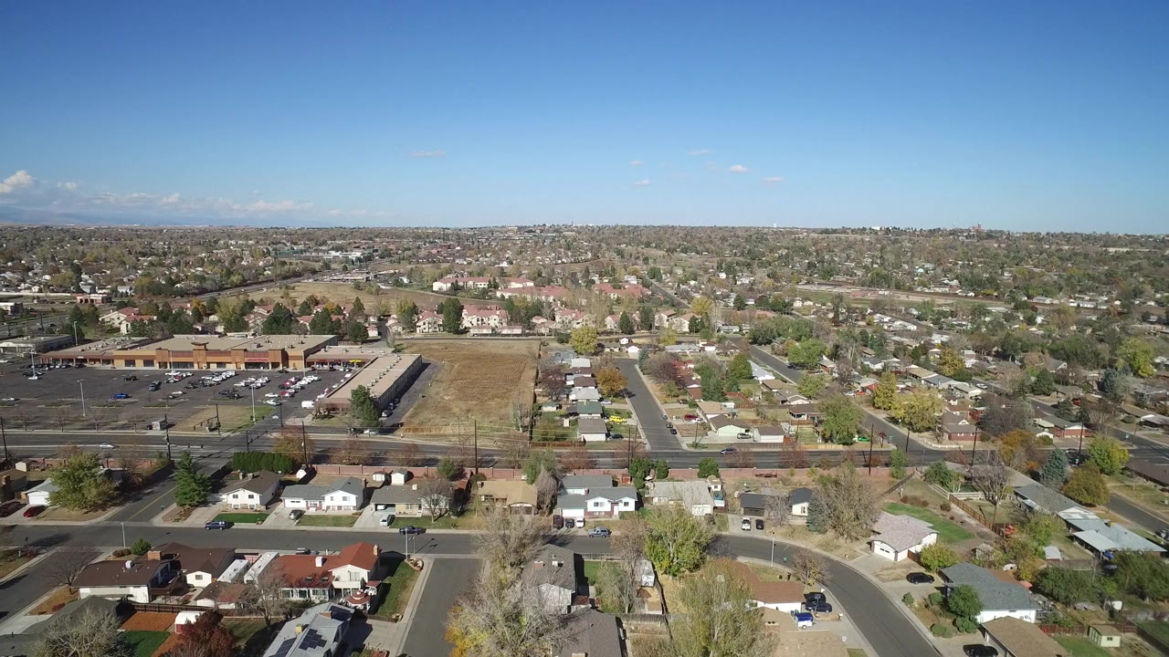 Aerial frame in Hidden Lake, Colorado, with a larger stretch of roadway and parking area visible at left, rows of homes across the center, dry open ground, and no visible people.