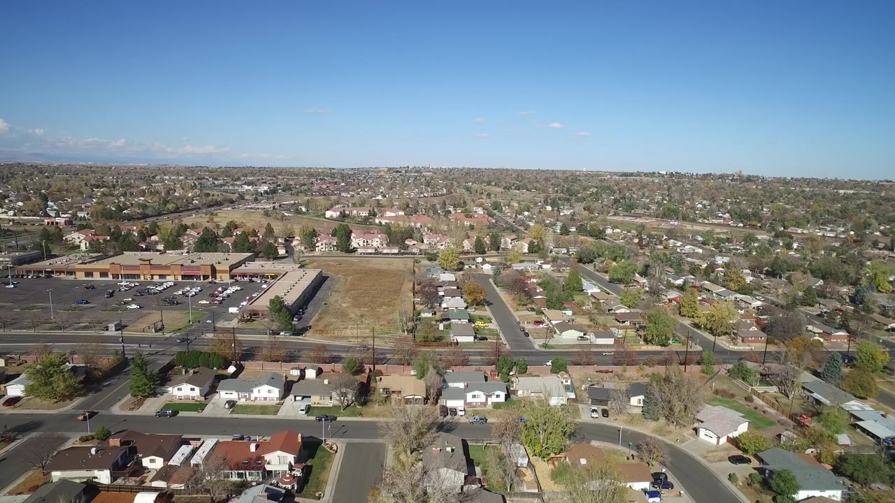 Second aerial frame in Hidden Lake, Colorado, showing the parking lot and long commercial-style building on the left, residential streets below, open ground in the middle, and no visible people.