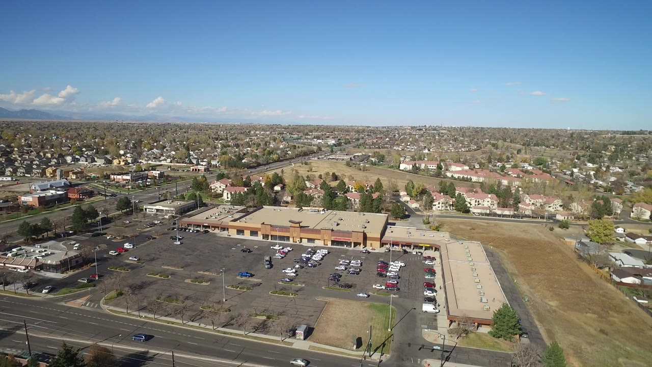 High aerial view of a shopping plaza, parking lots, roads, houses, trees, and distant mountains.