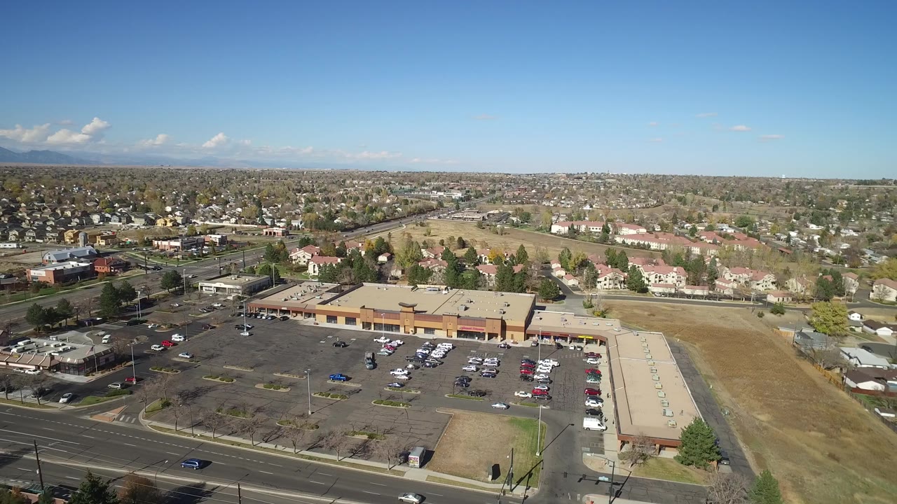 Hidden Lake, Colorado aerial frame centered on a tan-roofed commercial strip with parking areas below, roads at left and front, residential buildings beyond, open land, and no visible people.