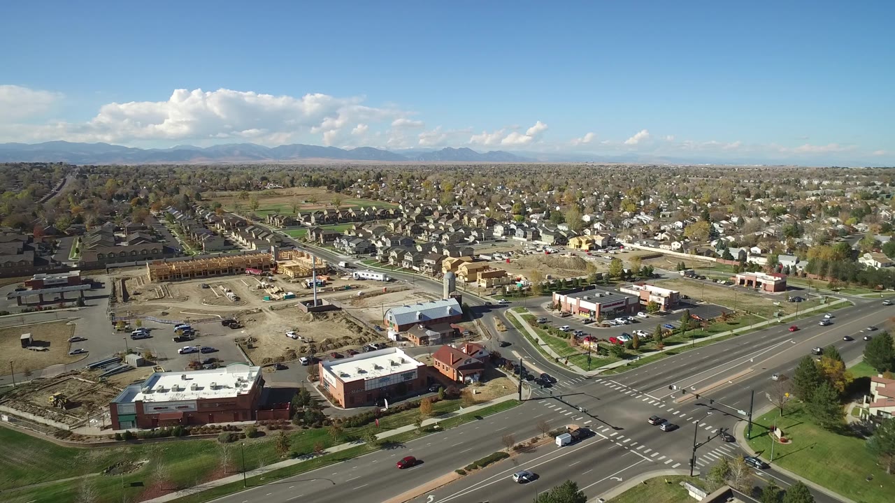 Aerial view in Hidden Lake, Colorado with a foreground intersection, construction lots, low retail buildings, dense houses, mountain horizon, and no visible people.