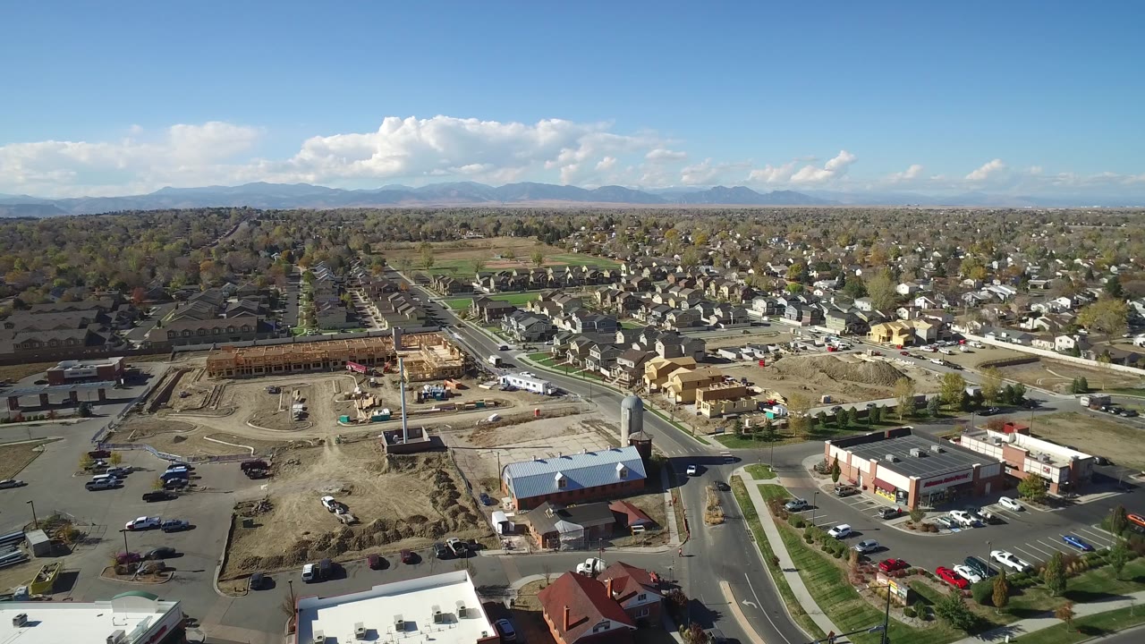 A high drone view in Hidden Lake, Colorado looks across a developed lot, nearby commercial buildings, residential streets, scattered vehicles, distant mountains, and no visible people.