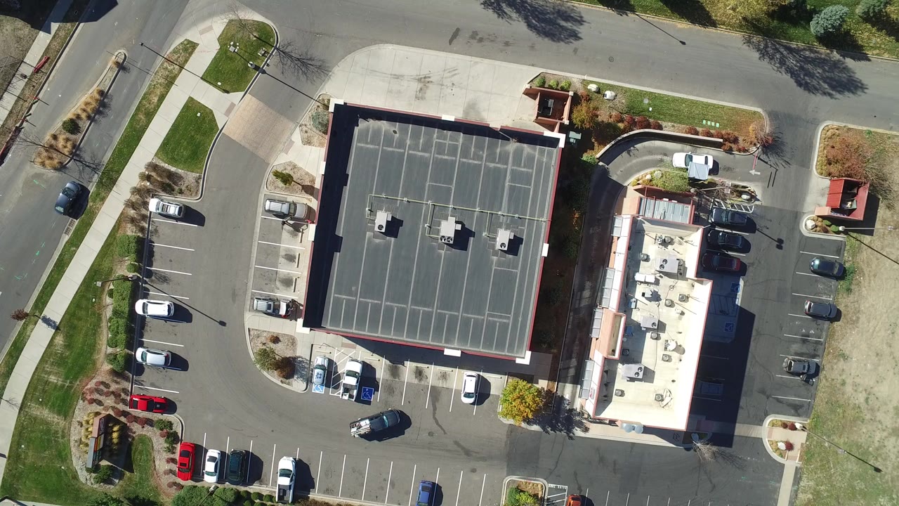 At 0:00, a vertical aerial frame over Hidden Lake, Colorado shows a dark flat-roofed building centered beside parking lots, roads, grass strips, and open dirt ground, with no visible people.