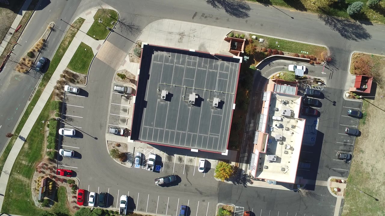 Vertical drone frame of a large roof, neighboring building, parking rows, and open ground beside a road.