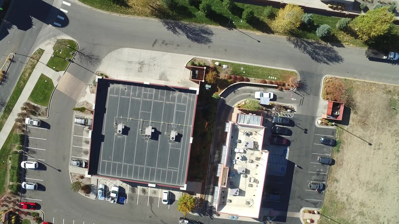Top-down aerial view of a flat-roof building, parking lot, road, trees, and dry grass lot.