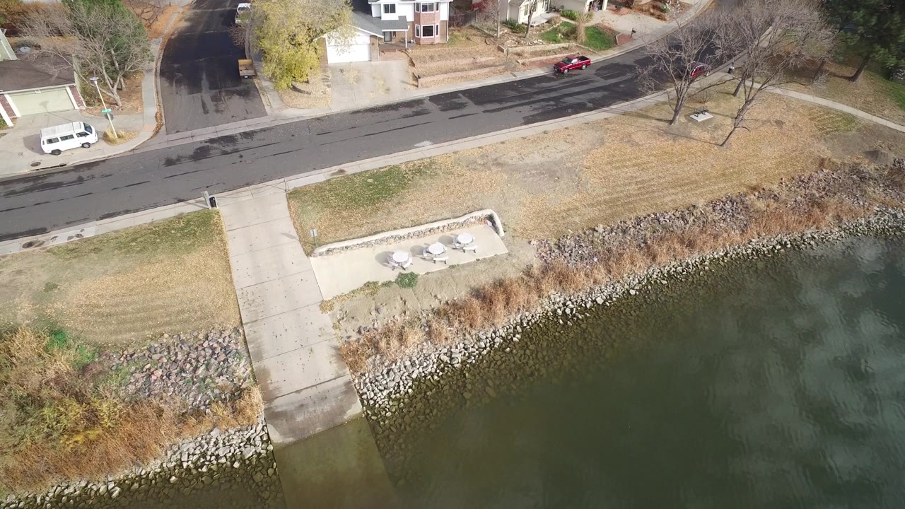 Aerial view over Hidden Lake, Colorado with the concrete ramp centered, green-blue water below, a road above the shoreline, residential lawns, houses, and no visible people.
