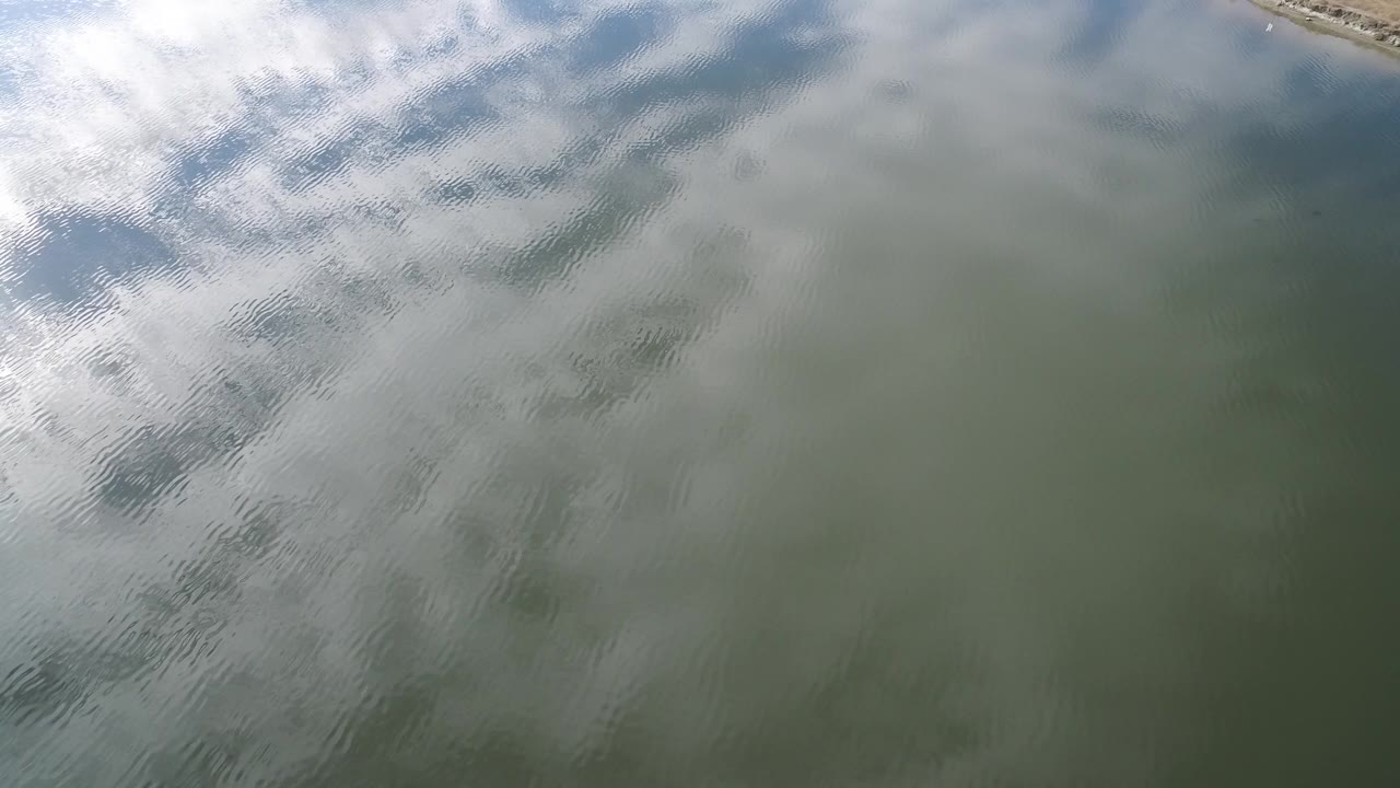Aerial frame over Hidden Lake, Colorado showing green-gray water, broken white cloud reflections across the upper half, darker ripple bands below, and no visible people.