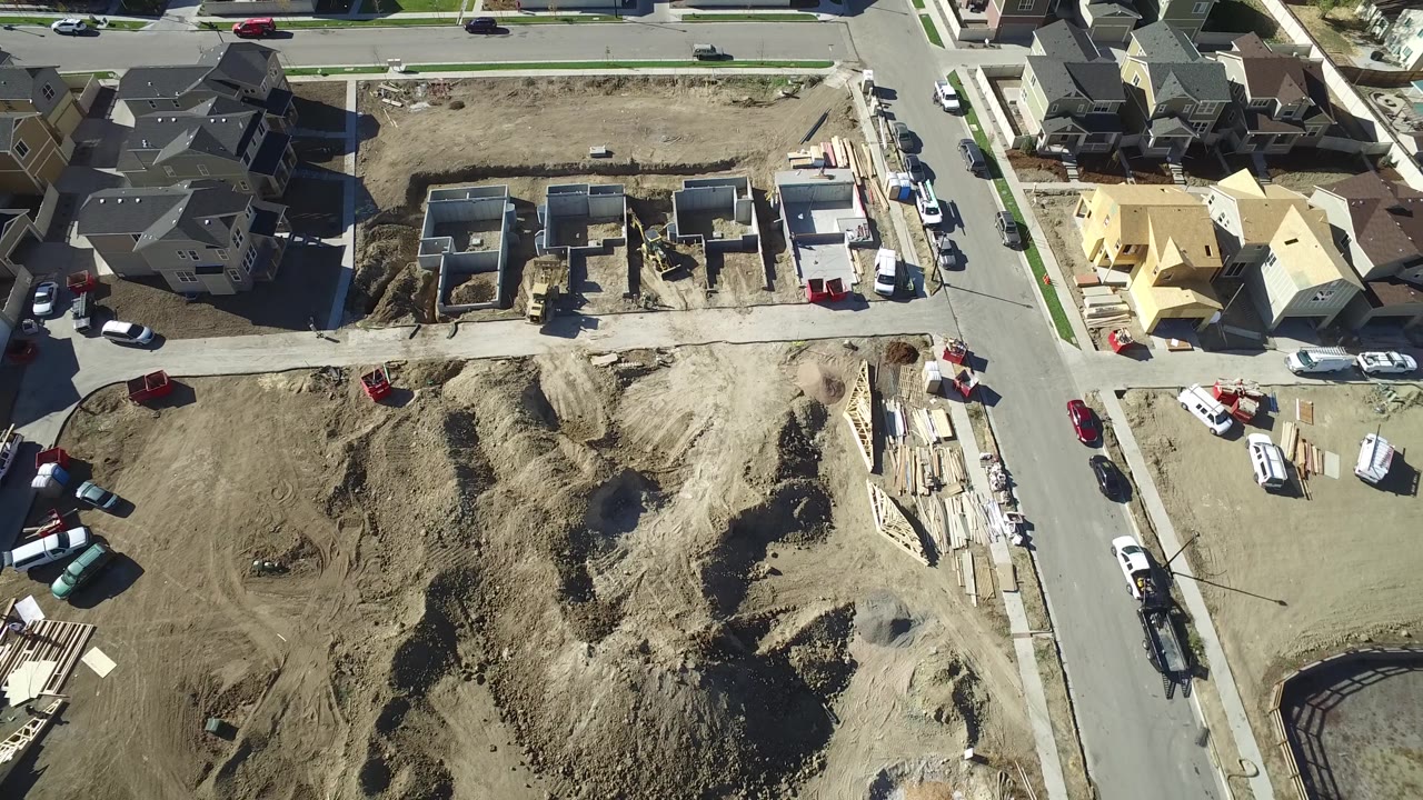 A high overhead frame in Hidden Lake, Colorado centers on a graded dirt lot and concrete foundations, with finished houses, streets, sidewalks, parked vehicles, and no visible people.