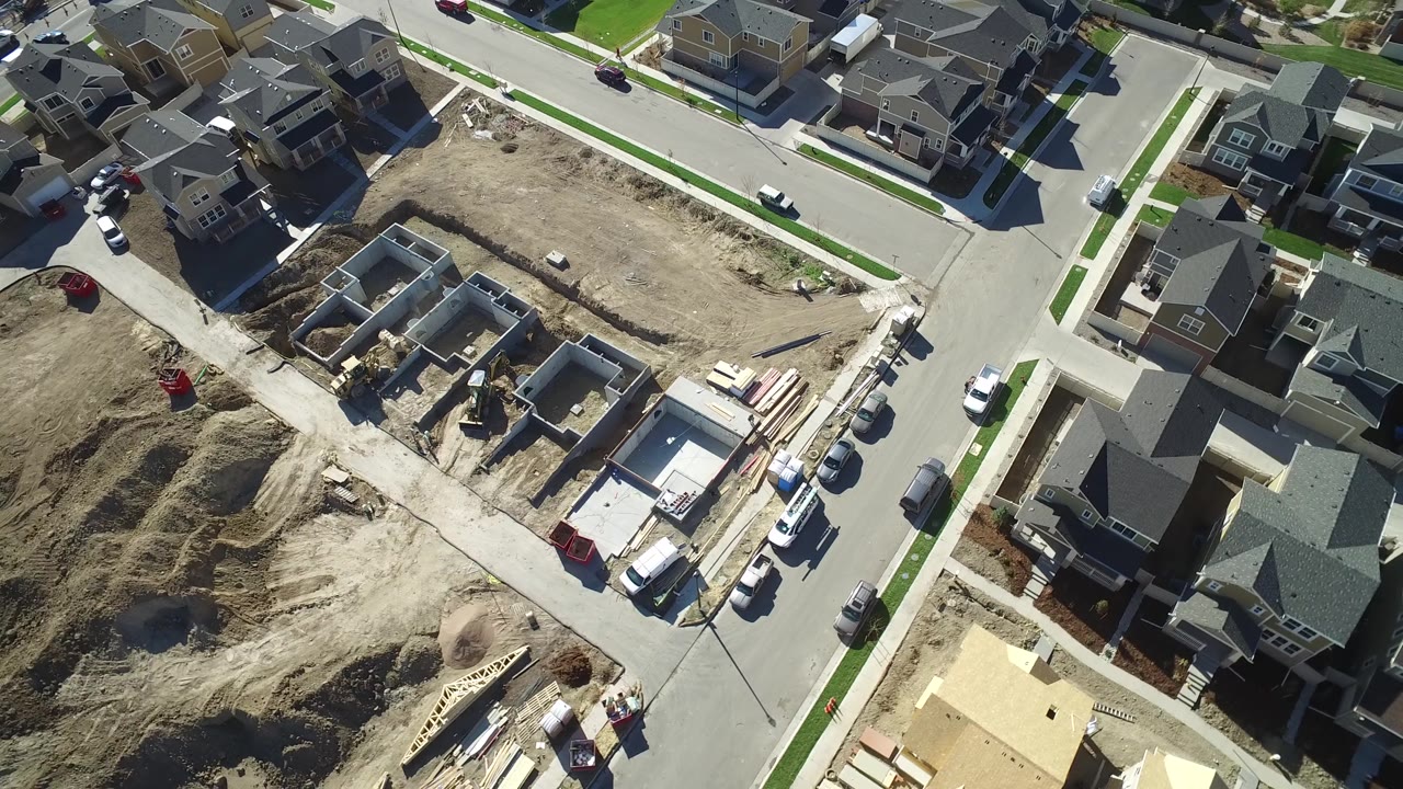 Aerial frame in Hidden Lake, Colorado with a partly built house foundation, stacked lumber, parked cars, nearby completed homes, and no visible people.