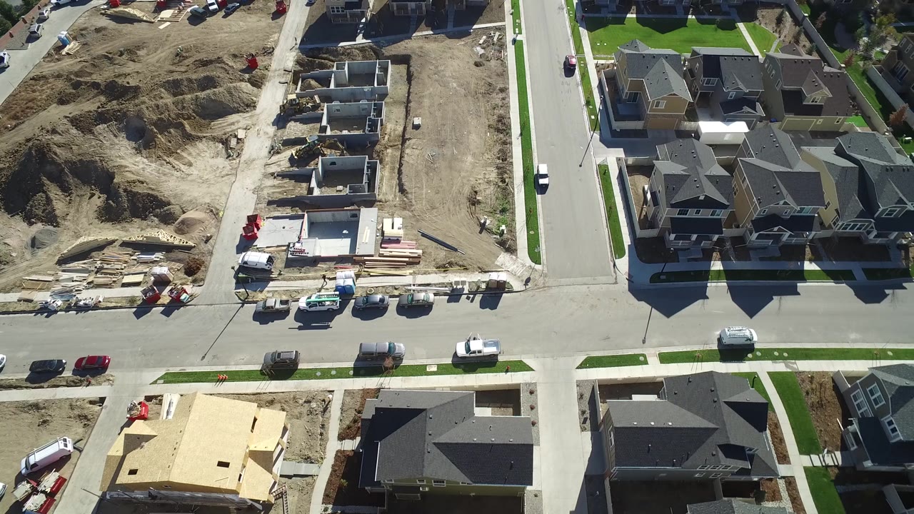 Aerial frame in Hidden Lake, Colorado with concrete foundations, excavated dirt, stacked building materials, finished houses across the street, and small possible workers too distant to identify.
