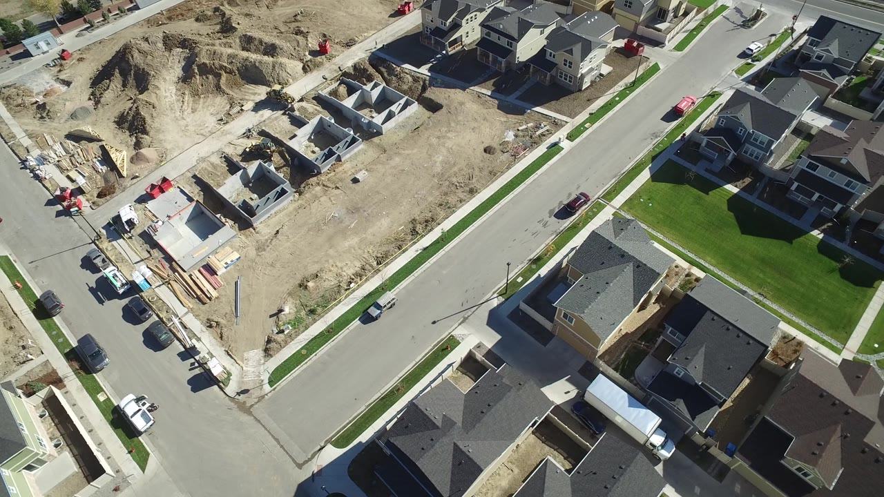 Aerial frame over Hidden Lake, Colorado, with a curved road crossing the view, completed homes on one side, construction lots on the other, parked cars, sidewalks, lawns, and no visible people.