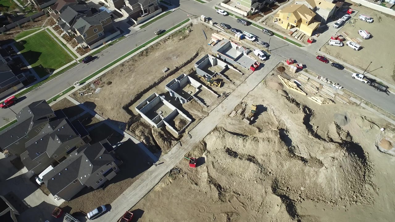 Drone-style overhead view in Hidden Lake, Colorado showing a broad dirt worksite, partially built home foundations, parked cars along the roads, nearby houses, and no visible people.
