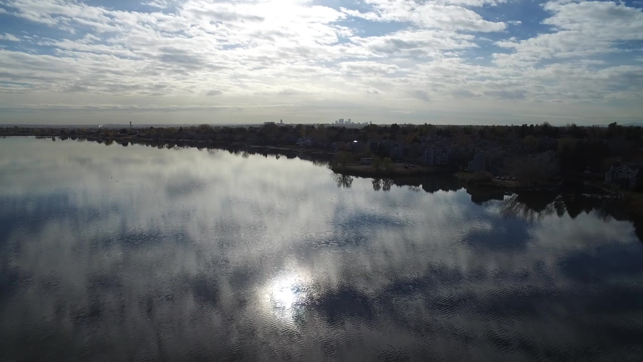 Wide lake surface reflecting clouds and sun beside a dark shoreline.