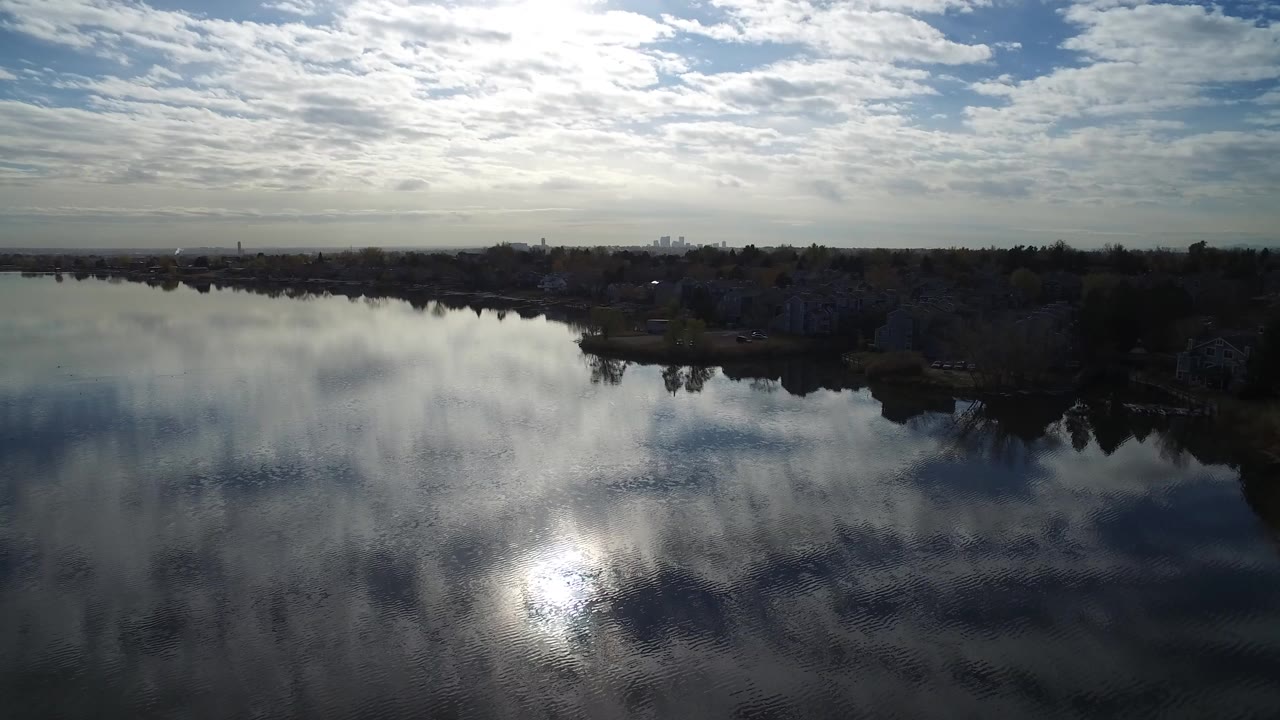Opening frame over Hidden Lake, Colorado, with bright sun reflected in the water, a dark shoreline, distant homes, and no visible people.
