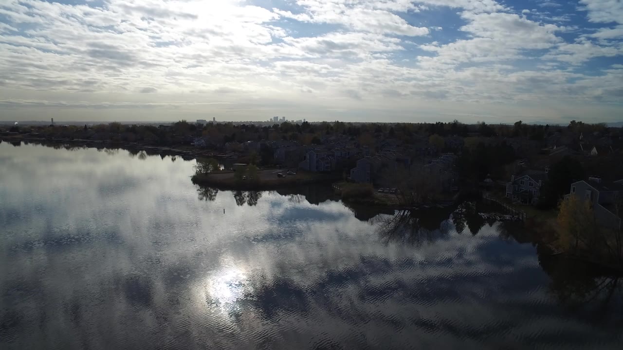 Aerial view over reflective lake water toward a dark shoreline and clouded sky.