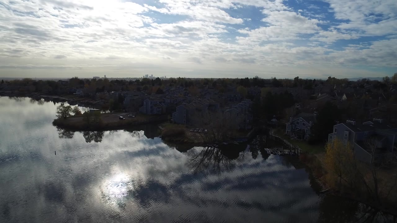 Aerial view over reflective water, trees, homes, and distant skyline beneath bright clouds.