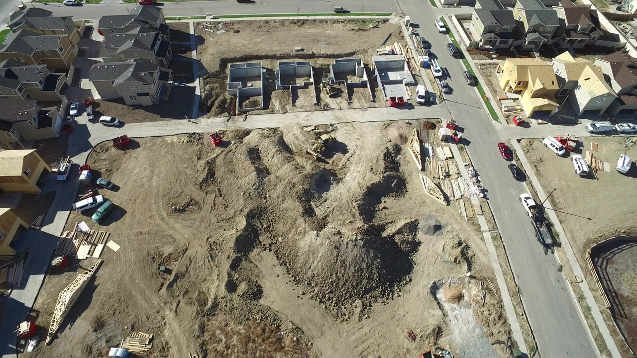 Overhead view in Hidden Lake, Colorado showing a dirt construction parcel, framed house sites, paved roads, parked vehicles, and nearby finished homes with no visible people.