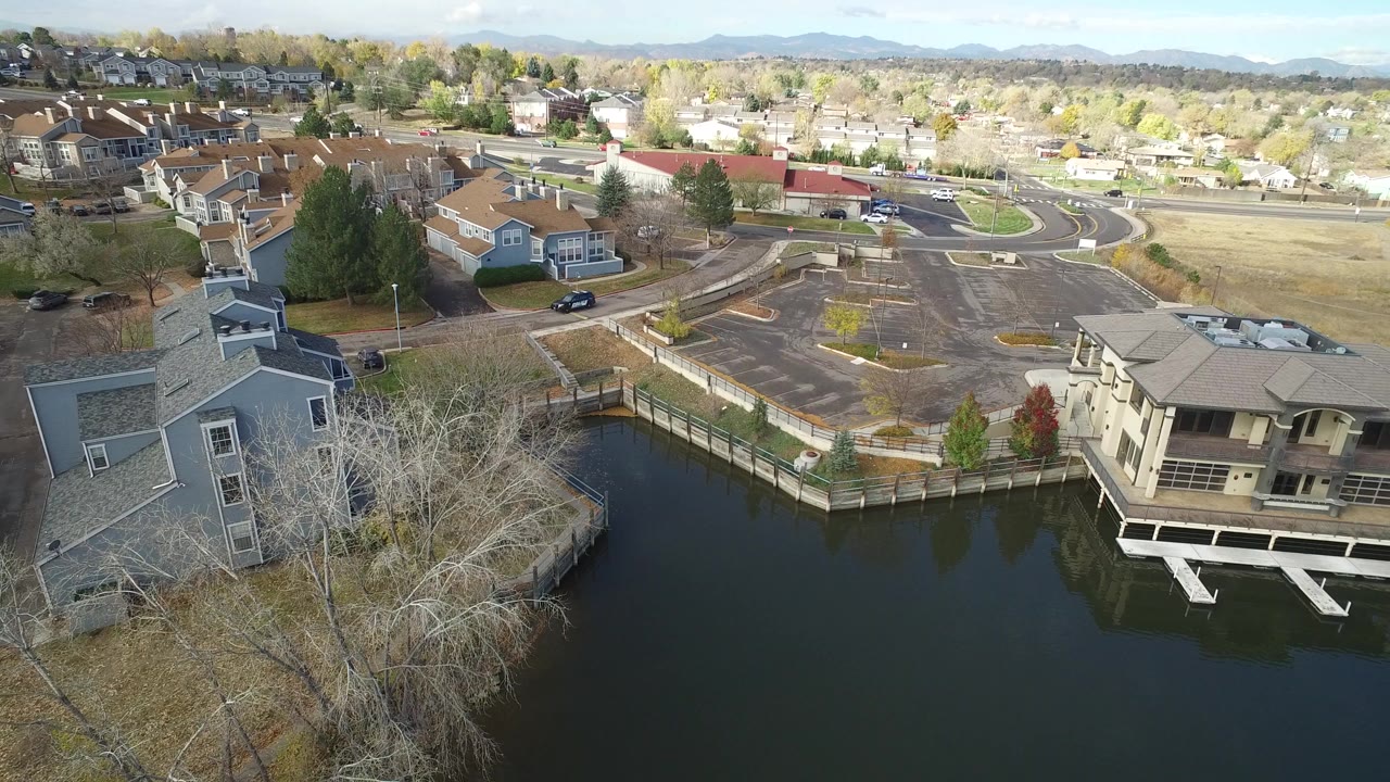 High aerial view over Hidden Lake, Colorado homes beside dark water, with curving roads, autumn trees, distant mountains, and no visible people.