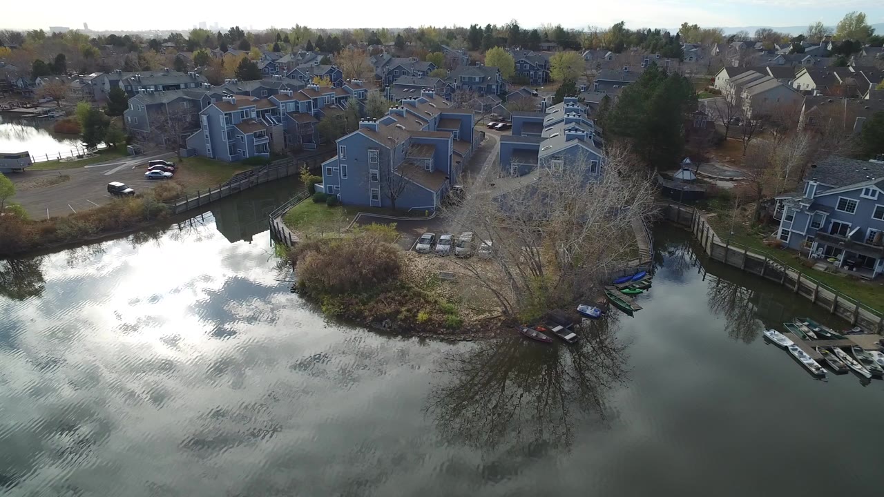 Aerial view over reflective lake water toward lakeside houses and small boats.
