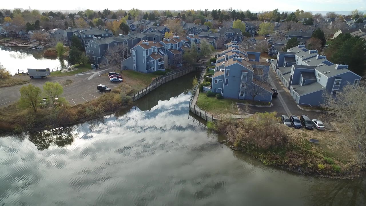 Aerial frame at Hidden Lake, Colorado, with the water channel centered below gray residential buildings, parked vehicles near the bank, and no visible people.