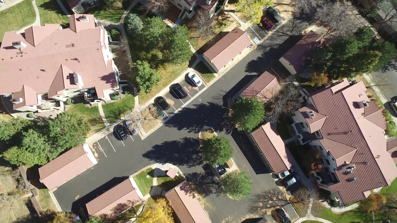 High overhead frame in Hidden Lake, Colorado showing clustered homes, an internal street junction, parking bays, lawns, trees, and hard midday shadows.