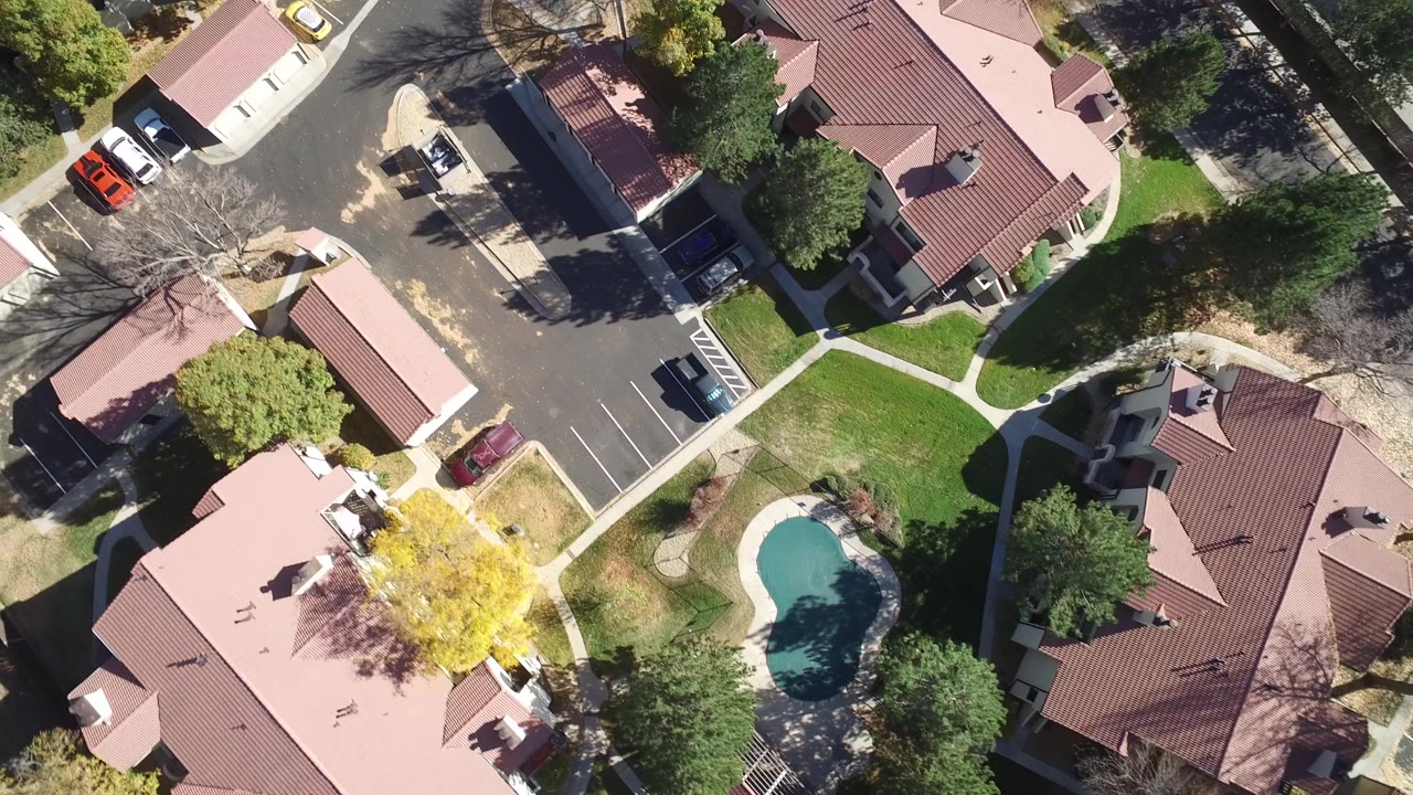 Aerial overhead frame in Hidden Lake, Colorado showing clustered red-roofed homes, a road bend, parked vehicles, sidewalks, lawns, trees, and the pool near the lower frame, with no visible people.