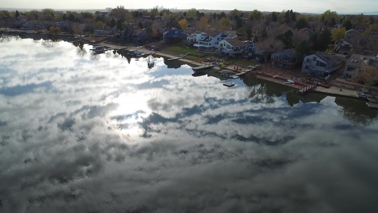 Aerial frame over Hidden Lake, Colorado, with bright cloud reflections on the water, lakeside homes along the upper shore, docks, trees, and small shoreline figures visible.