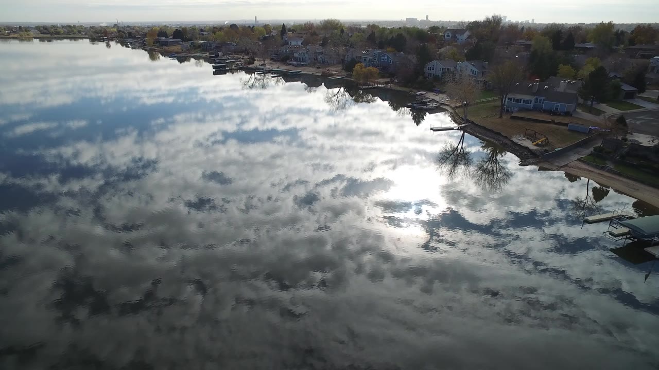 High aerial frame over Hidden Lake, Colorado, with cloud reflections across the water, shoreline homes at the top edge, docks or small waterfront structures, and no visible people.