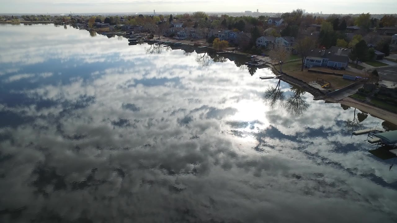Aerial view of Hidden Lake, Colorado, with bright sun reflection low on the water, a cluster of homes and leafless trees along the right shoreline, and no visible people.