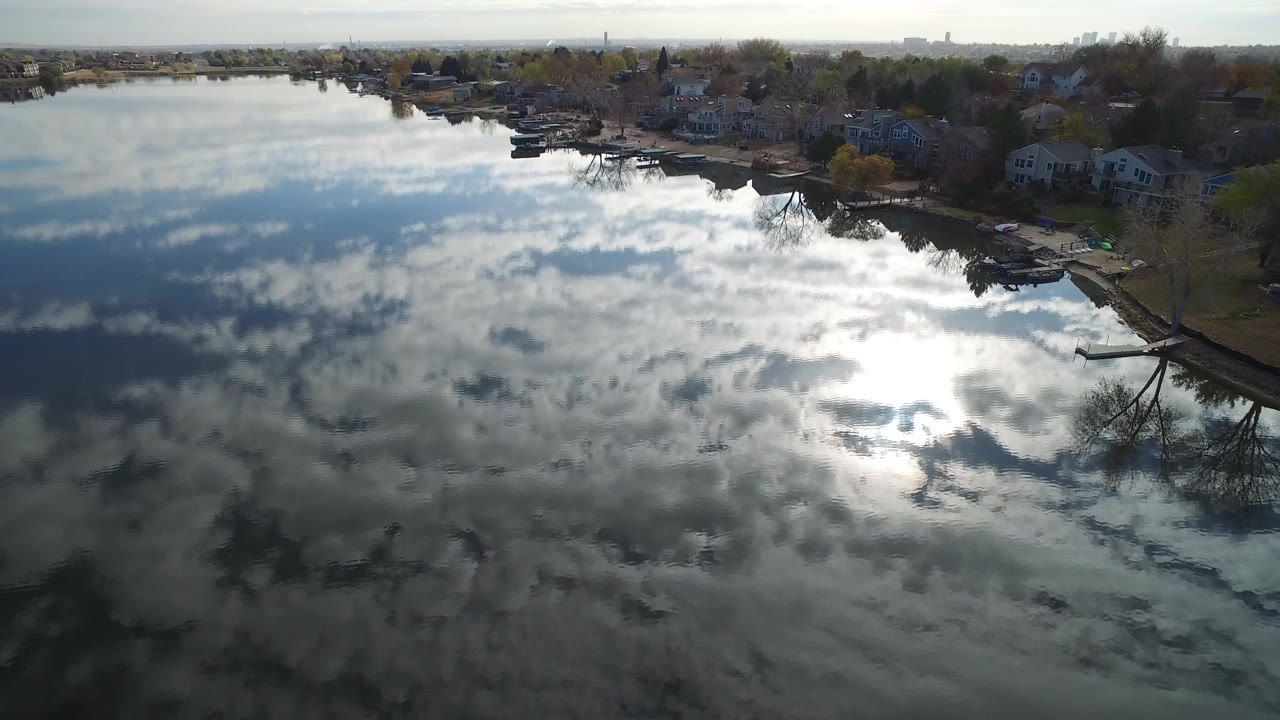 Aerial view over Hidden Lake in Colorado with cloud reflections on the water, shoreline houses and docks near the top of frame, and no visible people.