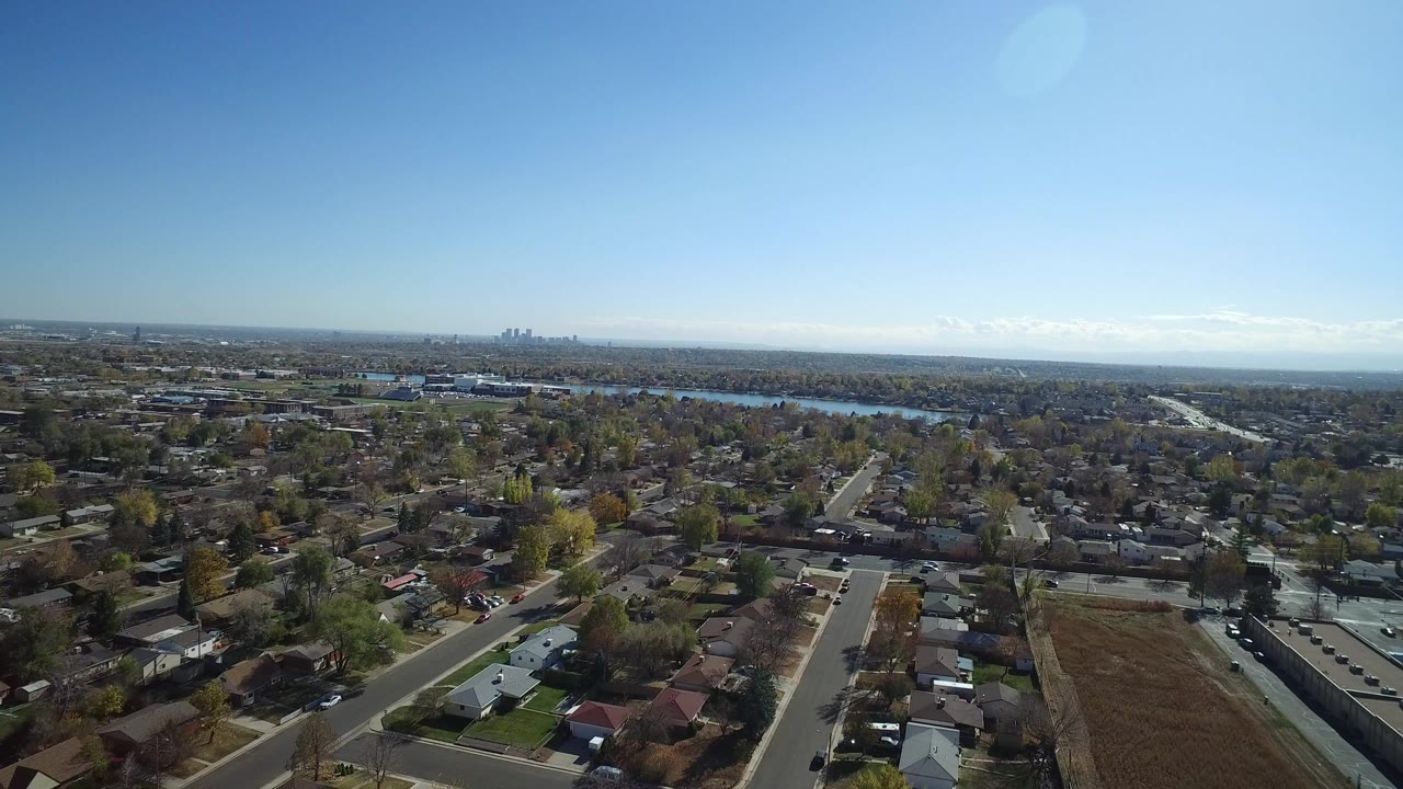 Aerial view over houses, streets, trees, open land, distant water, and skyline.