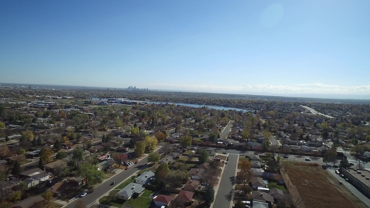 Aerial view over suburban streets, homes, trees, water, and distant skyline