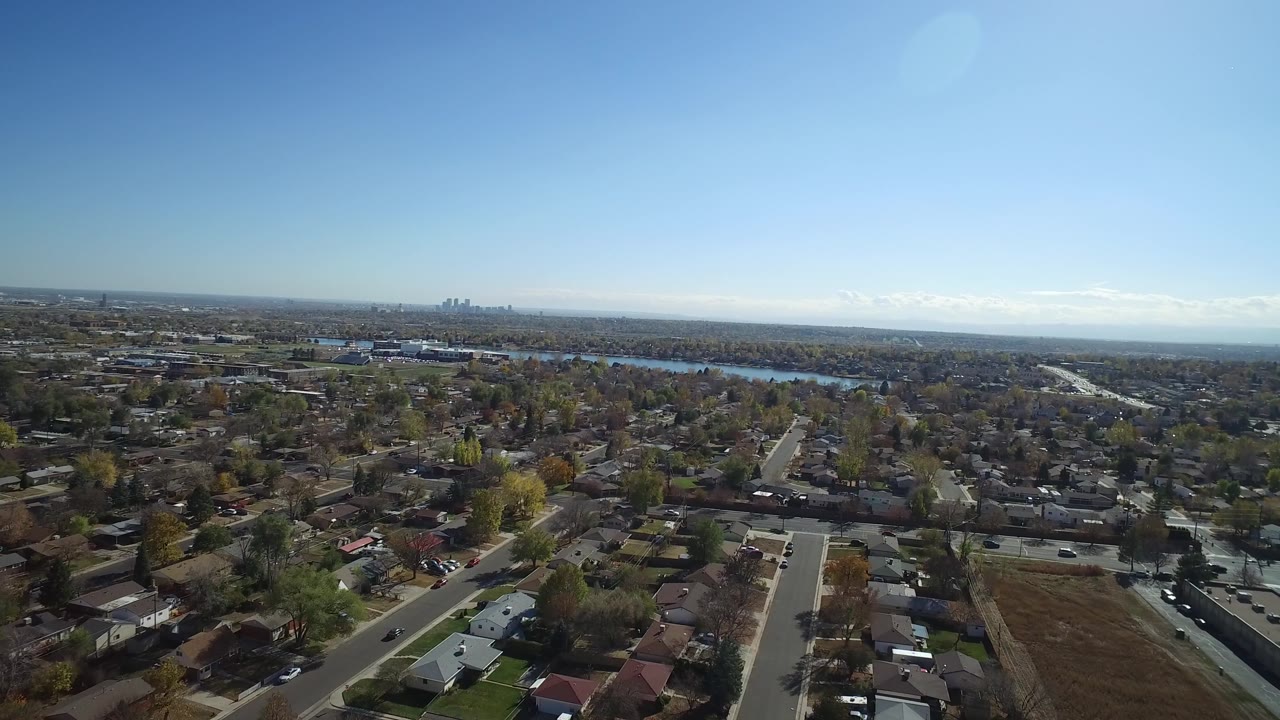 Aerial frame over Hidden Lake, Colorado, with a straight residential street leading toward tree cover, water in the midground, and a distant cluster of city buildings under clear sky.