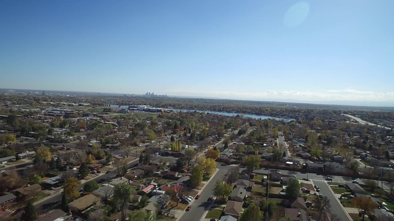 High aerial view over Hidden Lake, Colorado homes and tree-lined streets, with a blue water body and distant skyline under clear sky, no visible people.