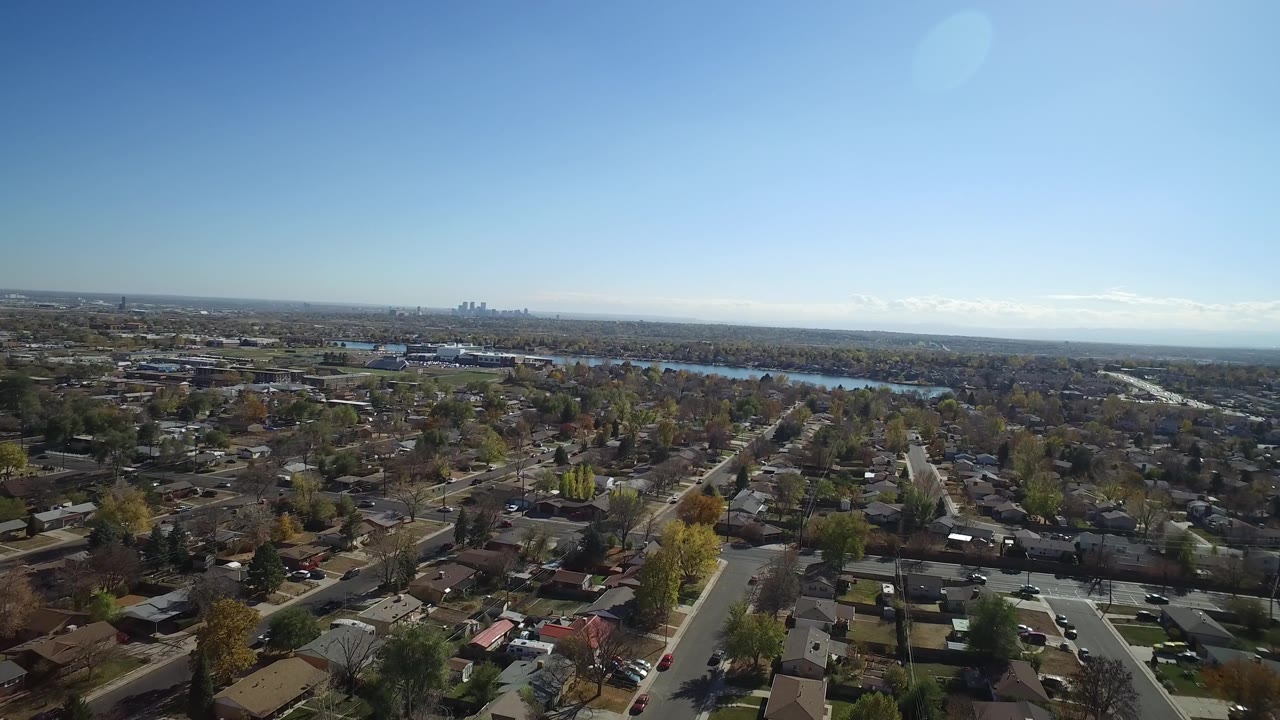 Wide aerial neighborhood view with rooftops, roads, distant lake, and skyline.