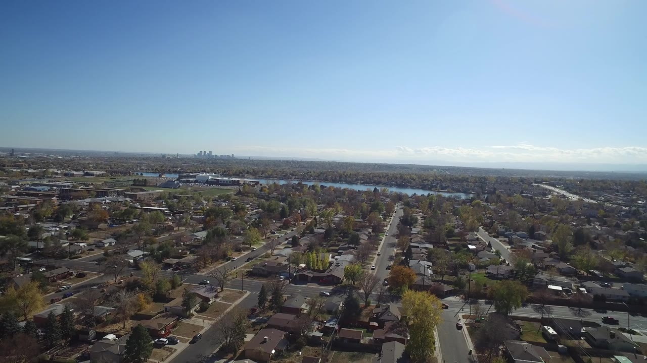 Aerial view of suburban houses, streets, trees, lake, and distant skyline under blue sky.