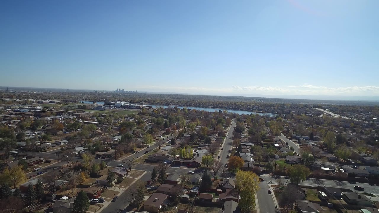 Aerial frame over Hidden Lake, Colorado, showing a central residential street, rooftops, scattered cars, the lake beyond the houses, distant buildings, and no visible people.